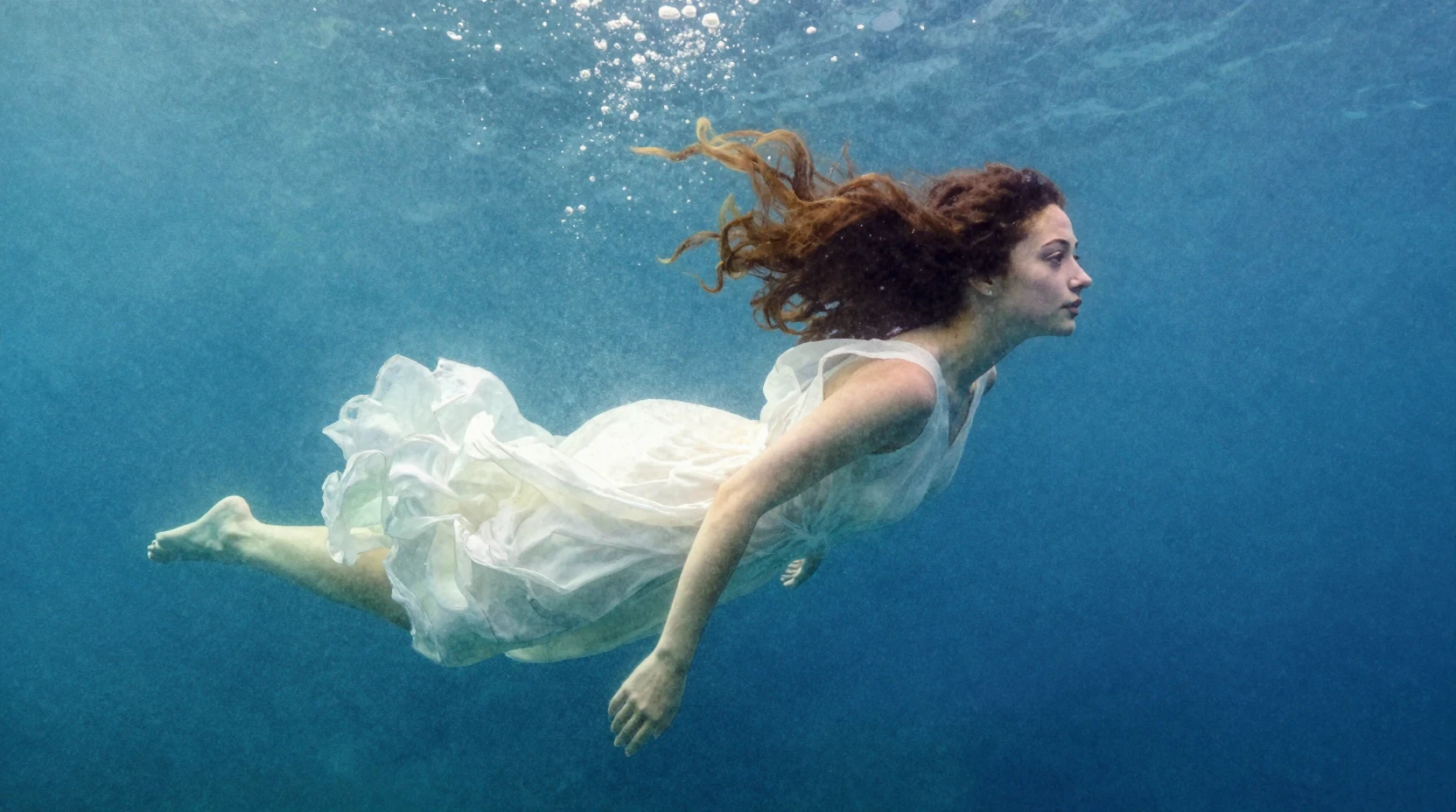 A woman in a white dress floats underwater with her curly red hair flowing