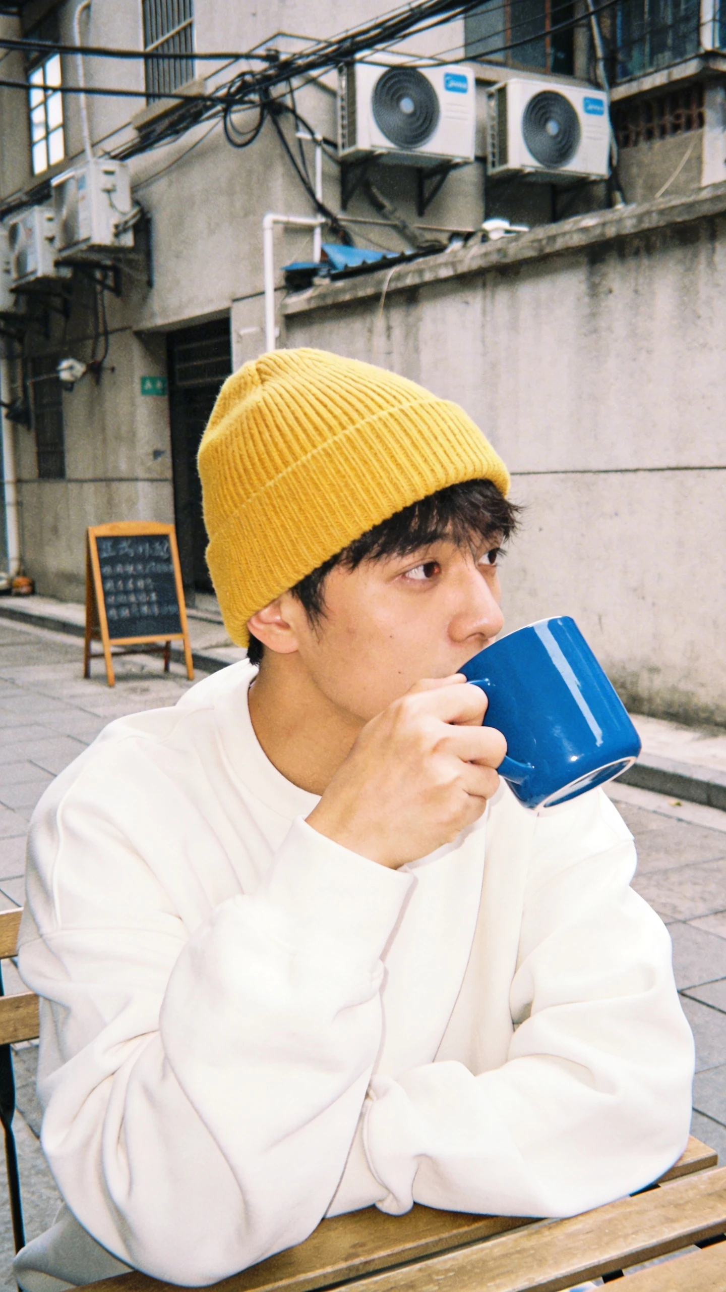 Young man in a yellow beanie and white sweatshirt drinking from a blue mug, in an alley.
