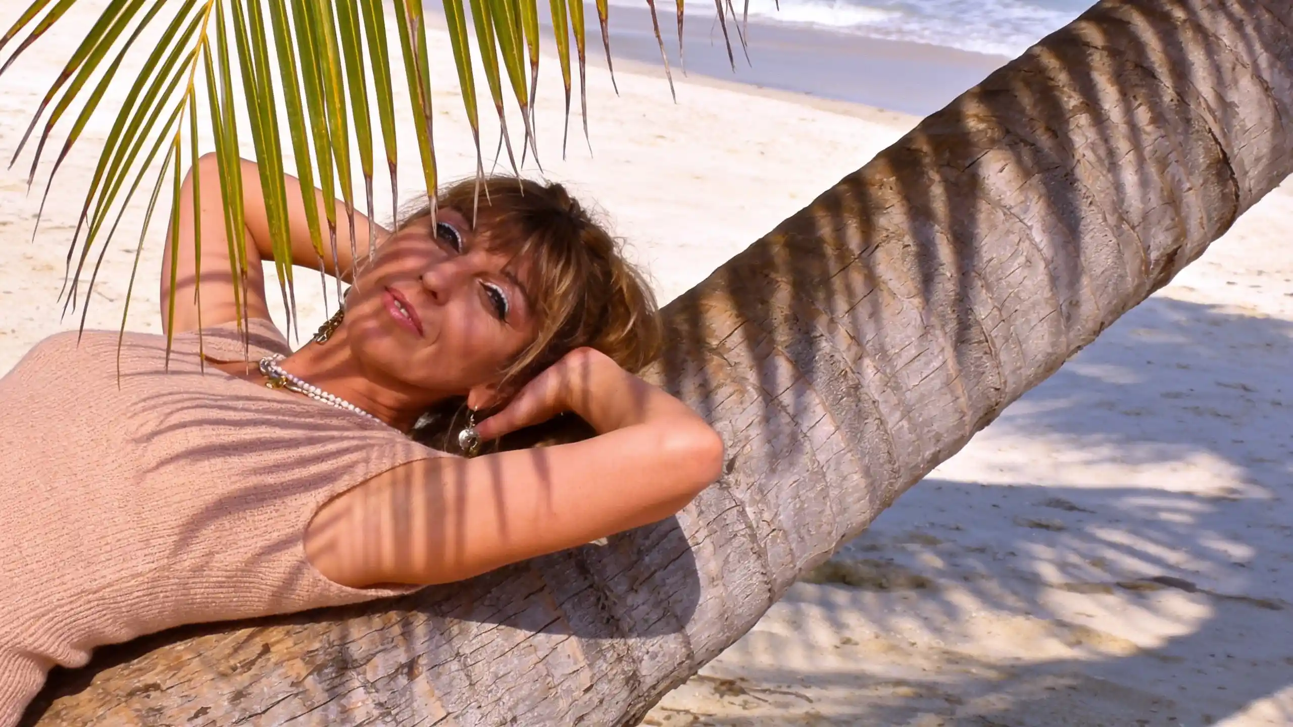 Blonde woman resting on a leaning palm tree by the beach