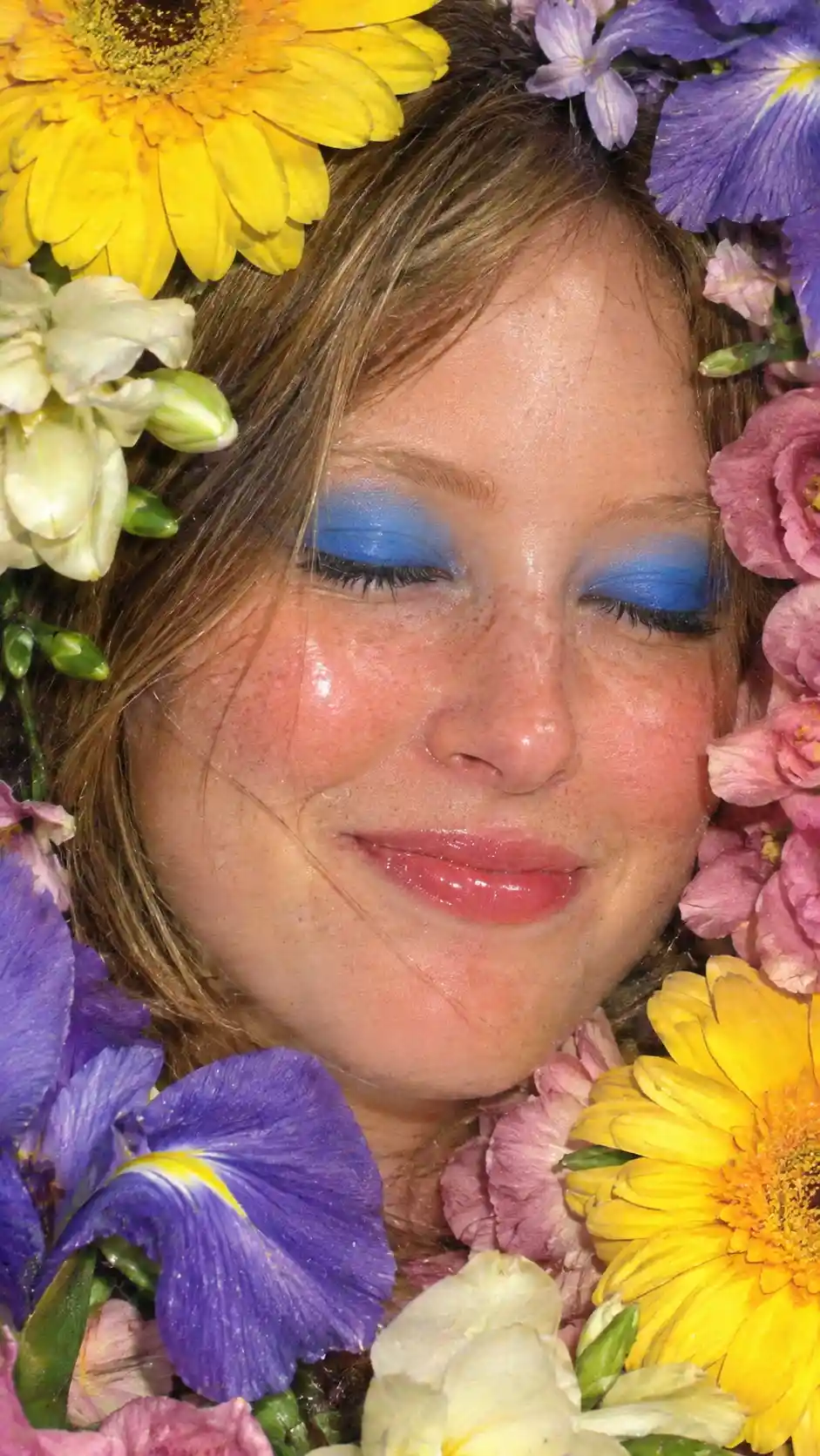 A young woman framed by vivid flowers and bold blue eyeshadow