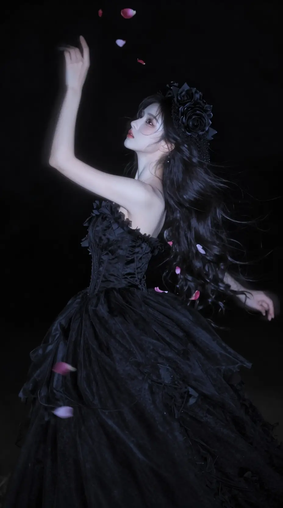 A woman rises from darkness in a black gown and rose fascinator