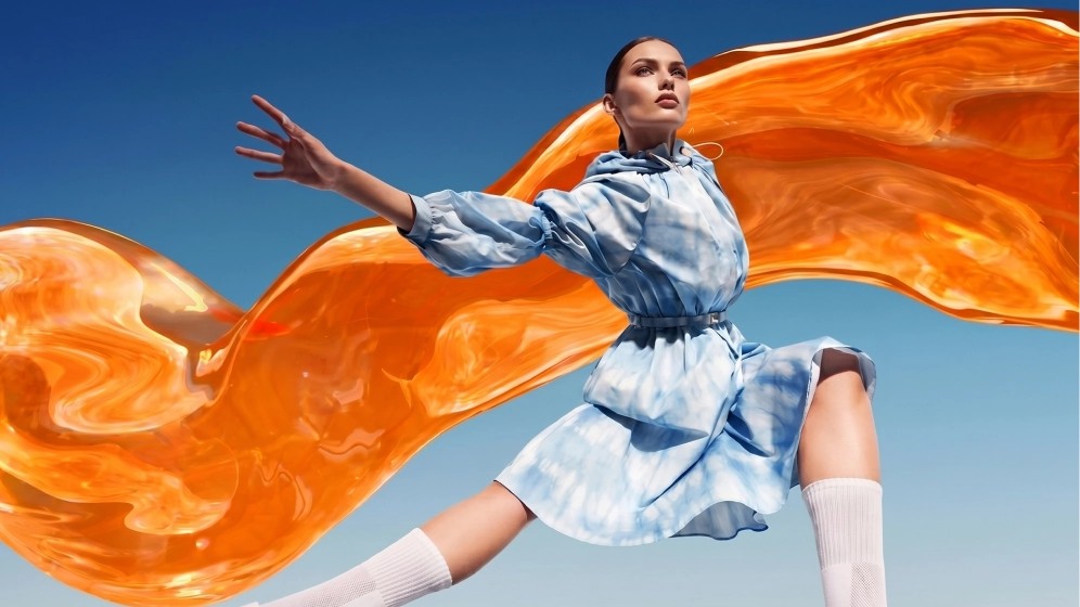 a woman in a blue tie-dye dress poses against a blue sky with a flowing orange liquid ribbon