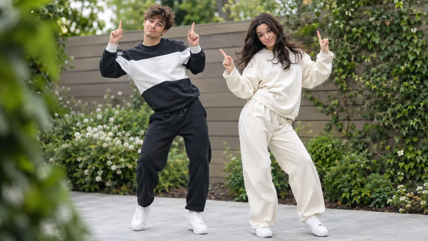 A young man and woman in casual sportswear dancing and pointing upwards in an outdoor garden setting.