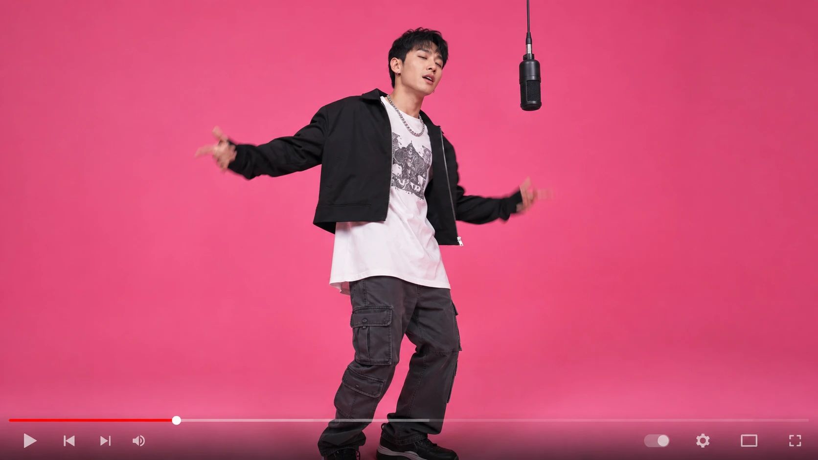 A young man dancing in a pink studio with a hanging microphone and video player controls.